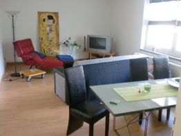 Wohnzimmer mit rotem Sessel, TV, Esstisch und Stühlen, helle Wände, Bild an der Wand.