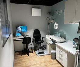 Kleine optometrieruimte met stoel, bureau, computer en oogmeetapparatuur.