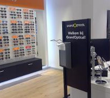 Winkelinterieur met een zonnebrillenwand en balie bij GrandOptical, met een Welkomstbord.