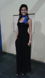 Uma mulher de vestido preto e lenço azul, sorrindo, com as mãos nos bolsos, posa em um local interno.