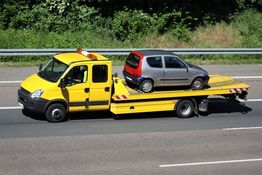 Gelbes Abschleppfahrzeug transportiert ein kleines graues Auto auf der Autobahn, umgeben von Bäumen.