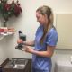 Smiling woman in scrubs holds a dental tool in a clinic room, with roses on a shelf nearby.