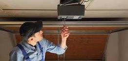 Man repairing a garage door opener mechanism in a garage while wearing a cap and overalls.