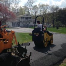 A person operates a roller to pave a driveway near a house surrounded by trees.