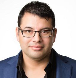 A man with glasses, dark hair, blue jacket, and black shirt smiles against a white background.