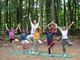 Eine Gruppe praktiziert Yoga im Wald, stehend im Baum-Pose auf Matten.