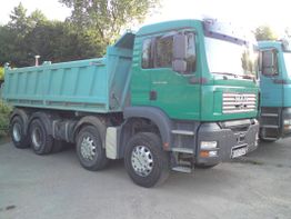 Grüner Lastwagen mit Kippmulde steht auf einem Parkplatz, umgeben von Bäumen.