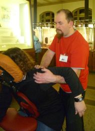 Mann in rotem T-Shirt massiert sitzende Person in einem öffentlichen Raum.