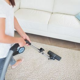 Person vacuuming a beige carpet in a living room with a white couch.
