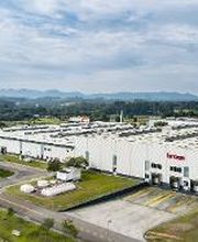 Fábrica grande em área verde, céu nublado ao fundo.