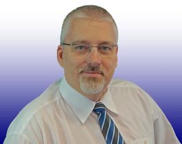 Ein Mann mit Brille und Bart trägt Hemd und Krawatte vor einem blau-weißen Farbverlauf.