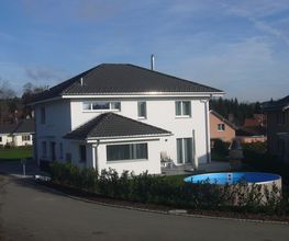 Zweistöckiges weißes Haus mit blauem Pool im Garten, umgeben von Hecke. Klarer Himmel im Hintergrund.