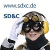 Person mit Schutzbrille und Gehörschutz, Text "www.sdxc.de" und "SD&C" im Hintergrund.