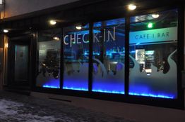 Außenansicht eines Cafés bei Nacht, mit neonblauer Beleuchtung und der Aufschrift "CHECK-IN".