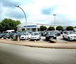 Autohandel mit verschiedenen Fahrzeugen auf dem Parkplatz, umgeben von Bäumen und Wolken.