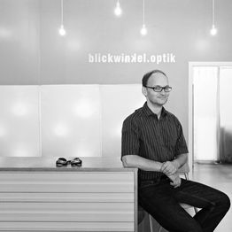 Mann in Optikergeschäft, lächelt und trägt Brille, auf einem Tisch liegt eine Sonnenbrille.