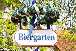 Schild "Biergarten" mit traditionellem Kranz, umgeben von frühlingshaften Bäumen.