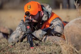 Hunter in camouflage and orange gear aims rifle, lying prone on grassy ground.