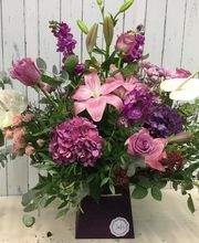 Ramo de flores variadas en tonos rosados y lilas sobre fondo de madera blanca.