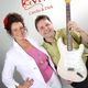 Zwei Personen posieren fröhlich mit einer E-Gitarre vor einem Logo mit der Aufschrift "Revival".