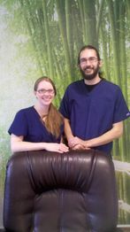 Zwei Menschen in blauer Kleidung lächeln hinter einem Sessel vor einer Tapete mit Bambusmotiv.