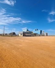 Estrada de terra com edifício industrial ao fundo e caminhões estacionados, sob céu azul.