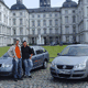 Zwei Personen stehen vor zwei Autos vor einem großen Gebäude im Hintergrund.