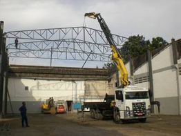 Caminhão com guindaste ergue estrutura metálica em construção. Funcionários ao redor observam.