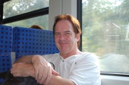 Ein Mann lächelt im Zug und lehnt sich an den Sitz. Draußen ist eine grüne Landschaft zu sehen.
