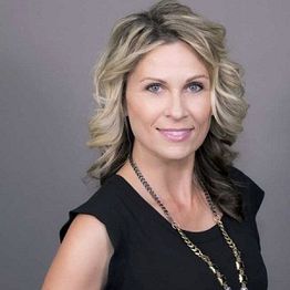 Smiling woman with wavy hair in a black top, wearing a necklace, against a gray background.