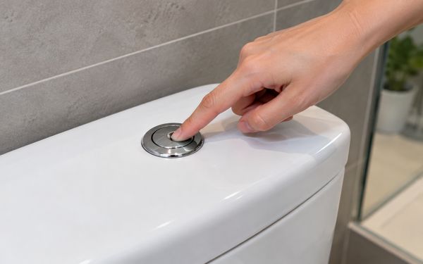 Nahaufnahme einer Hand, die den Spülknopf eines weißen Toilettenspülkastens betätigt.