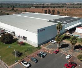 Vista aérea de una gran nave industrial blanca con estacionamiento y palmeras alrededor.