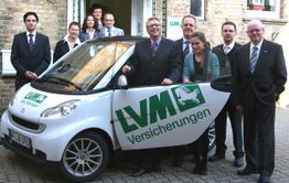 Menschen stehen neben einem Smart-Auto mit LVM Versicherungen Logo vor einem Backsteingebäude.