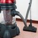 Vacuum cleaner with red top and clear canister on carpeted floor near a wall.
