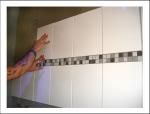 Hände platzieren weiße Fliesen mit grauem Mosaik-Band an der Wand.
