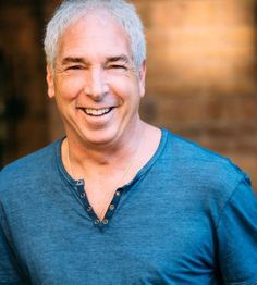Smiling man in a blue shirt standing against a blurred, warm-toned background.