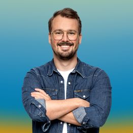 Ein Mann mit Brille und Bart lächelt und verschränkt die Arme vor einem blauen Hintergrund.