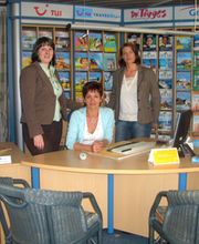 Drei Personen in einem Reisebüro mit Prospekten im Hintergrund, eine sitzt am Schreibtisch.