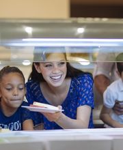 Familia sonríe mientras escoge comida en un buffet, adultos ayudan a niños.