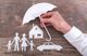 Hand holding umbrella over paper family, house, and car cutouts on wooden background.