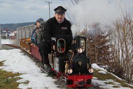Mann fährt dampfende Miniaturlokomotive mit Passagieren durch verschneite Landschaft.