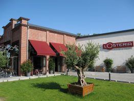 Rote Backsteingebäude von L'Osteria mit Terrasse und grünem Baum im Vordergrund.