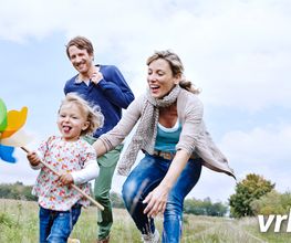 Familie spielt im Freien, Kind mit Windrad, Eltern lachen, blauer Himmel im Hintergrund.