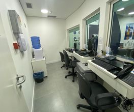 Sala de escritório com mesas, cadeiras, computadores e bebedouro ao fundo.