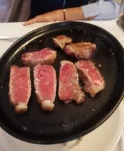 Carne asada sobre plancha negra, junto a copa de vino tinto y tabla de cortar en mesa blanca.