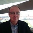 En mand med briller og jakke står foran et vindue med udsigt over en by og en flod.