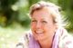 Ältere Frau lacht im Freien bei sonnigem Wetter, umgeben von unscharfem Grün im Hintergrund.