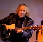 Ein Mann spielt eine Akustikgitarre, trägt eine dunkle Lederjacke vor einem neutralen Hintergrund.