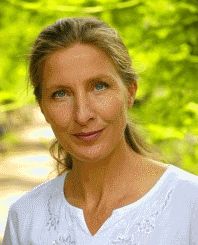 Frau in weißem Oberteil lächelt im Freien mit grüner Natur im Hintergrund.
