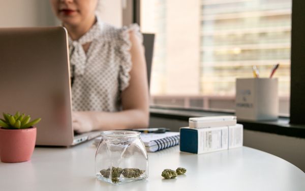 Cannabisblüten in einem Glas auf einem Schreibtisch neben Medikamentenpackungen, während eine Frau am Laptop arbeitet.
